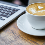 The image of a cup of cappuccino next to a laptop. In the foreground are the logos of the HPC Café Erlangen and BayernKI.