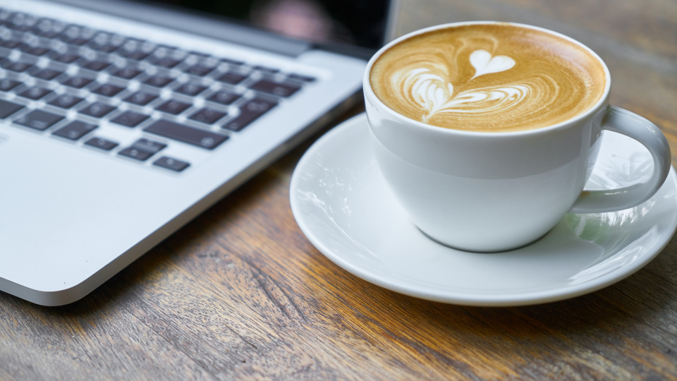 The image of a cup of cappuccino next to a laptop. In the foreground are the logos of the HPC Café Erlangen and BayernKI.