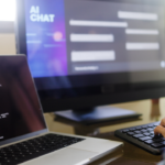 The picture shows a desk and the hands of a person working on two different computers. One is a desktop PC showing an AI chat program, the other is a laptop on displaying code.
